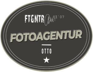 Fotograf Goslar, Logo Fotoagentur Otto, Hochzeitsfotograf. Logo der Fotoagentur Otto: Ein ovales, graues Emblem mit einem dreifachen Rand in unterschiedlichen Grautönen. Im oberen Bereich steht in einer weißen, abgenutzten Schrift 'FTGNTR' und daneben 'SI '07'. In der Mitte befindet sich der Schriftzug 'FOTOAGENTUR' in großen, schattierten Buchstaben. Darunter ist ein horizontaler Strich und darunter steht 'OTTO' in weißer Schrift. Am unteren Rand des Logos ist ein weißer Stern abgebildet.