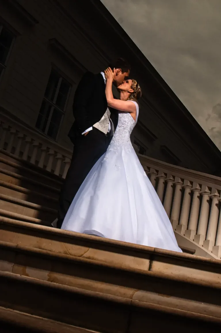 Eine Braut im weißen Kleid und ein Bräutigam im schwarzen Anzug küssen sich auf einer eleganten Treppe vor einem Gebäude unter einem dramatischen, wolkigen Himmel. Die Szene, wunderschön eingefangen von dem Fotografen aus Goslar und Deiner Region, ist romantisch und die Beleuchtung stimmungsvoll.