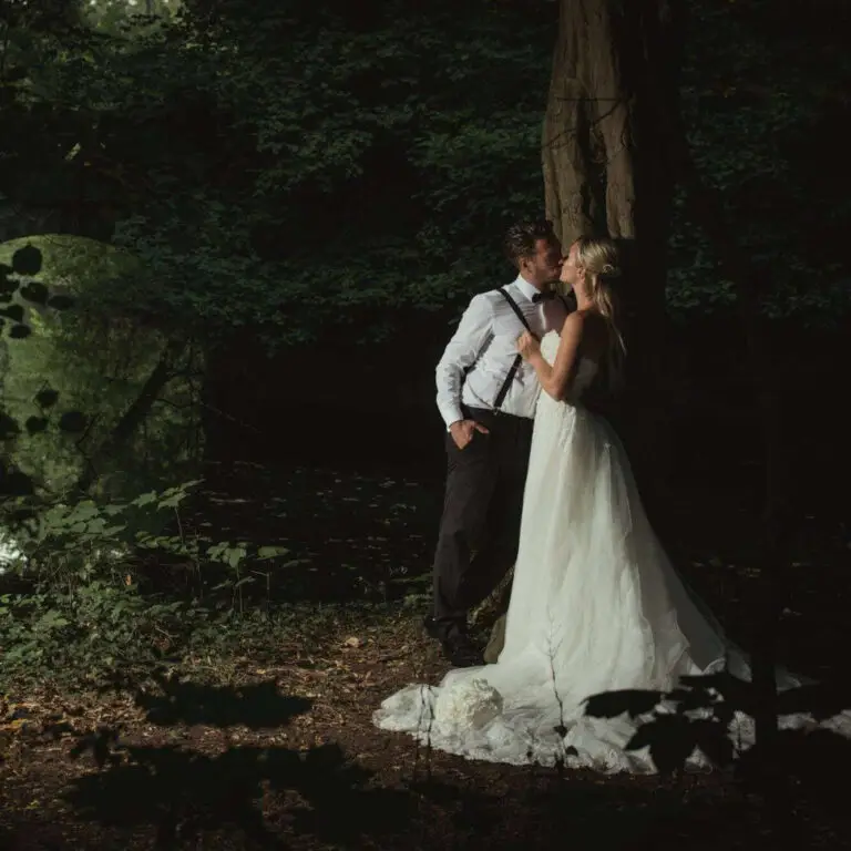 Ein romantisches Hochzeitsfoto, aufgenommen von Fotograf Goslar und Fotoagentur Otto, zeigt ein Brautpaar in einem dunklen Wald. Die Braut trägt ein elegantes weißes Kleid, während der Bräutigam schwarze Hosen, ein weißes Hemd und Hosenträger trägt. Sie stehen eng beieinander und schauen sich liebevoll an. Im Hintergrund ist ein Wasserlauf und ein Brückenbogen sichtbar, was dem Bild eine geheimnisvolle und romantische Atmosphäre verleiht.